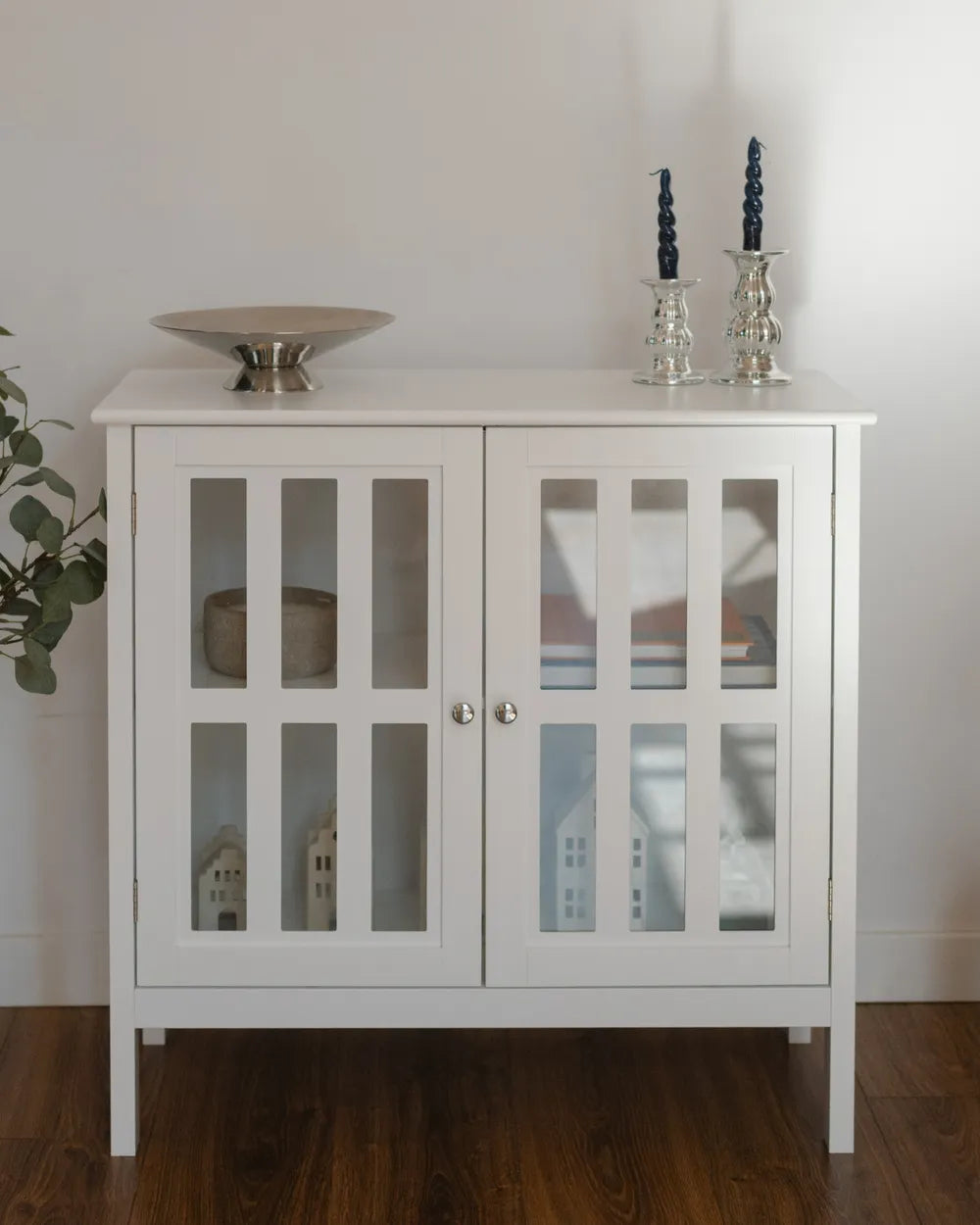 A white wooden sideboard cabinet with two glass-paneled doors, styled with decorative objects on top, set against a plain white wall and dark wood floor.