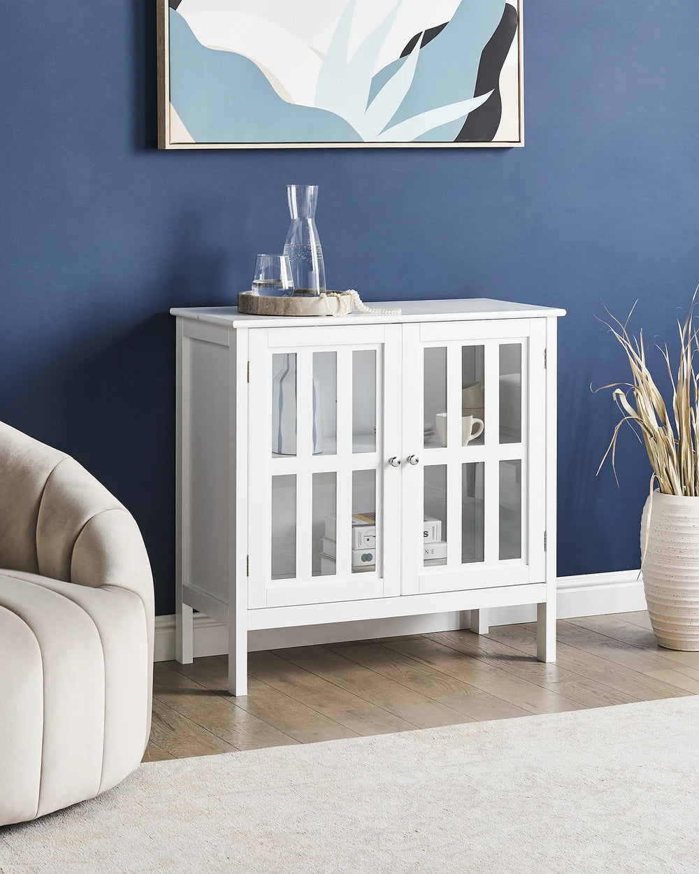 A white wooden sideboard cabinet with two glass-paneled doors stands against a dark blue wall in a styled room. Decorative items sit on top, and a beige armchair and dried plant arrangement are partially visible.