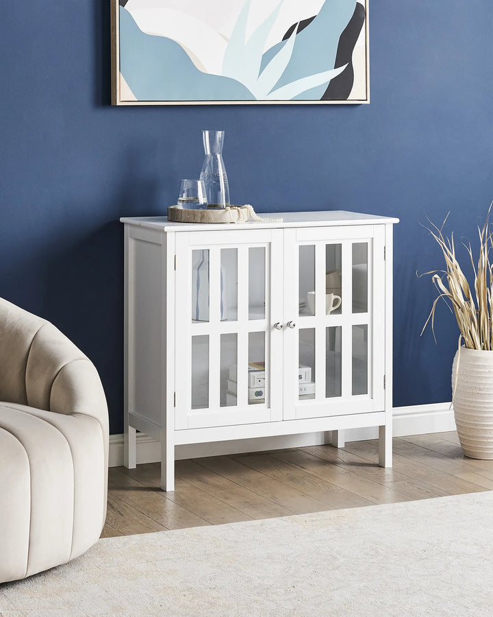 A white wooden sideboard cabinet with two glass-paneled doors stands against a dark blue wall in a styled room. Decorative items sit on top, and a beige armchair and dried plant arrangement are partially visible.