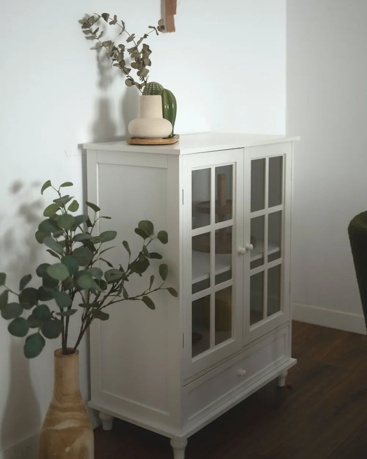 A white wooden cabinet with two glass-paneled doors and a single drawer, styled in a home setting with potted plants and a vase on top, set against a plain white wall and dark wood floor.