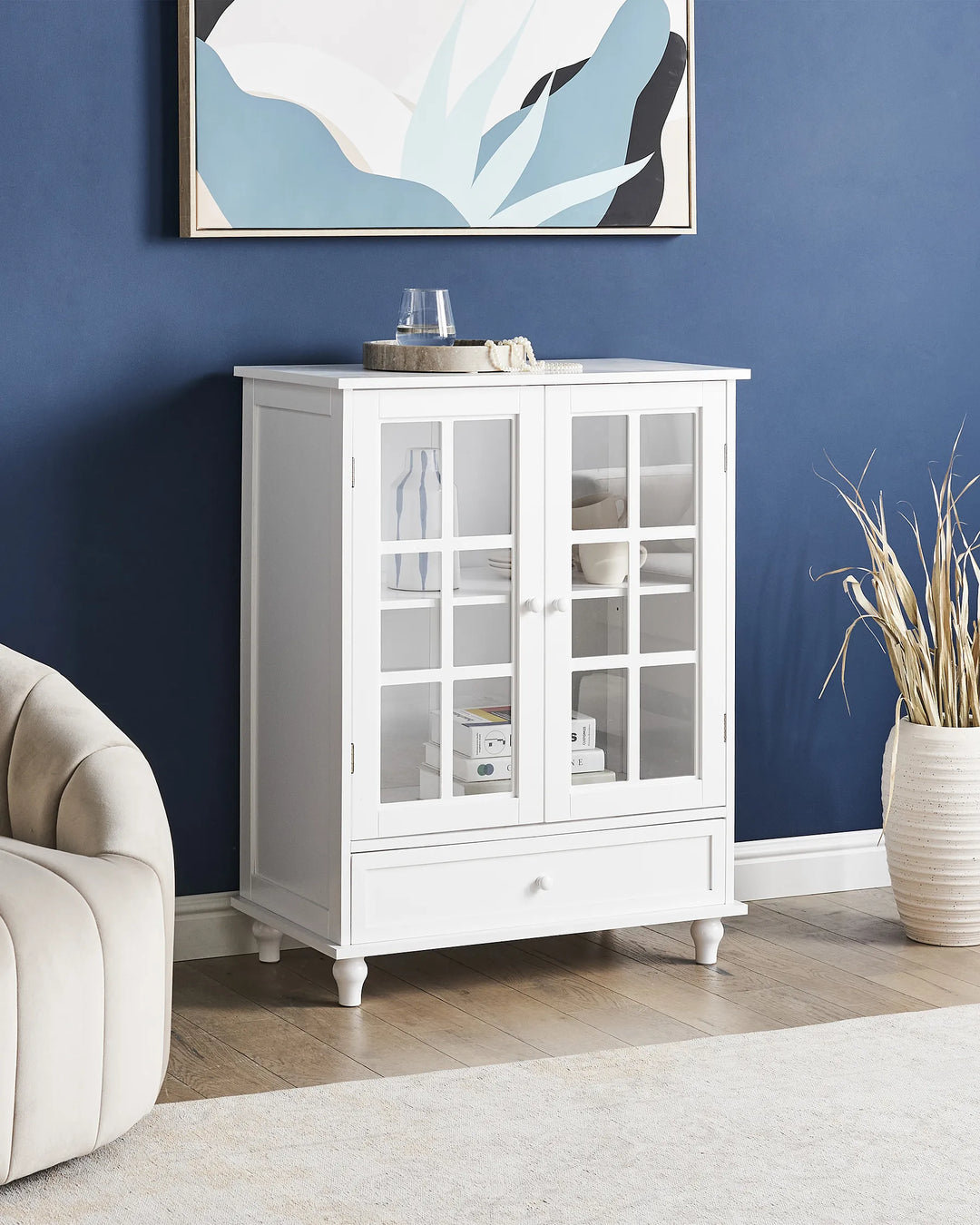White cabinet with glass doors in a room with blue walls and a white armchair.