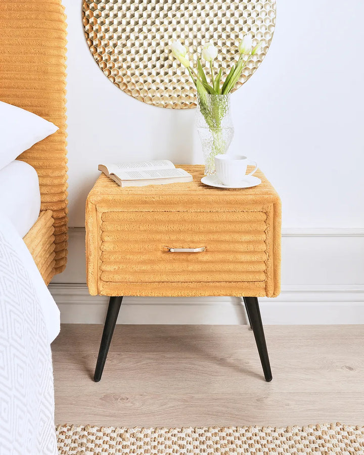 A mustard yellow textured fabric nightstand with black tapered legs, styled in a bedroom next to a bed with a woven headboard. The nightstand features a single drawer with a small metallic handle and is decorated with a vase of white tulips, an open book, and a white coffee cup.
