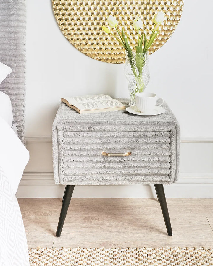 A light gray ribbed fabric nightstand with black tapered legs, styled in a bedroom setting with a white bed, a vase of tulips, an open book, and a white cup on top.