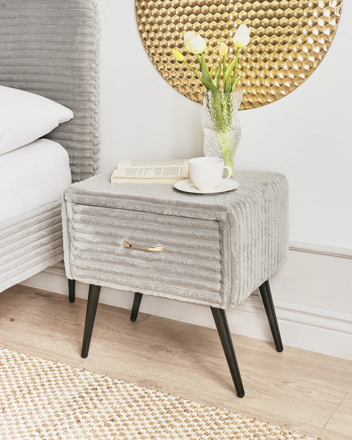 Gray upholstered side table with a white cup and saucer, and a vase of flowers in a room with a textured wall.