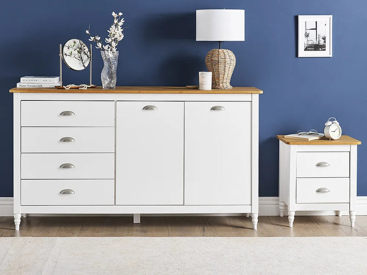A white wooden sideboard or dresser with six drawers and two cabinet doors, topped with a light natural wood surface. It is styled in a bedroom setting against a dark blue wall, alongside a matching nightstand, a lamp, and decorative items.