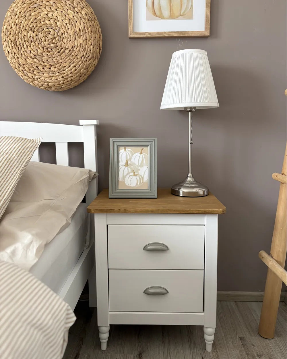 A white two-drawer nightstand with a light wood top, styled in a bedroom next to a bed with beige bedding. On top of the nightstand sits a white pleated table lamp with a silver base and a small framed picture.