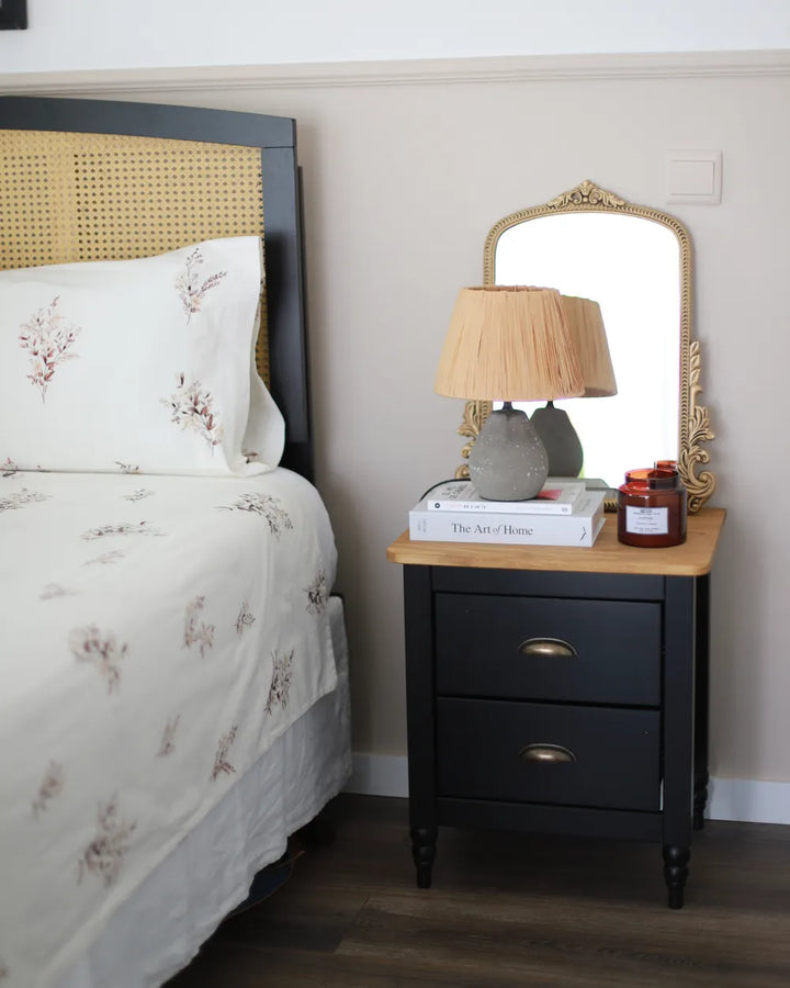 A styled bedroom corner featuring a black nightstand with a light wood top, an ornate gold-framed mirror, a beige lamp, and books, next to a bed with white floral bedding and a woven cane headboard.