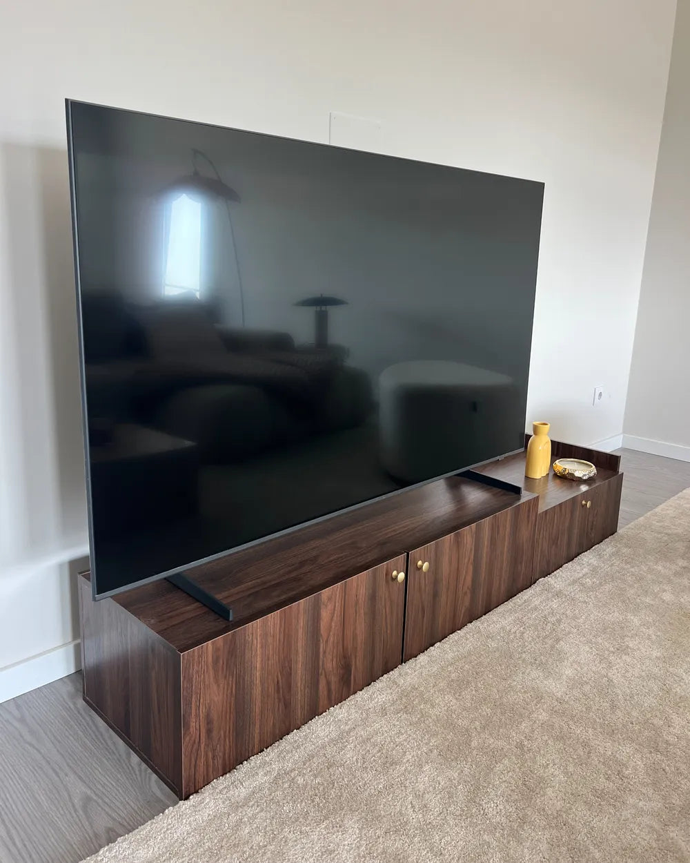A large flat-screen television mounted above a dark wood media console in a home interior with light walls and a beige carpet.