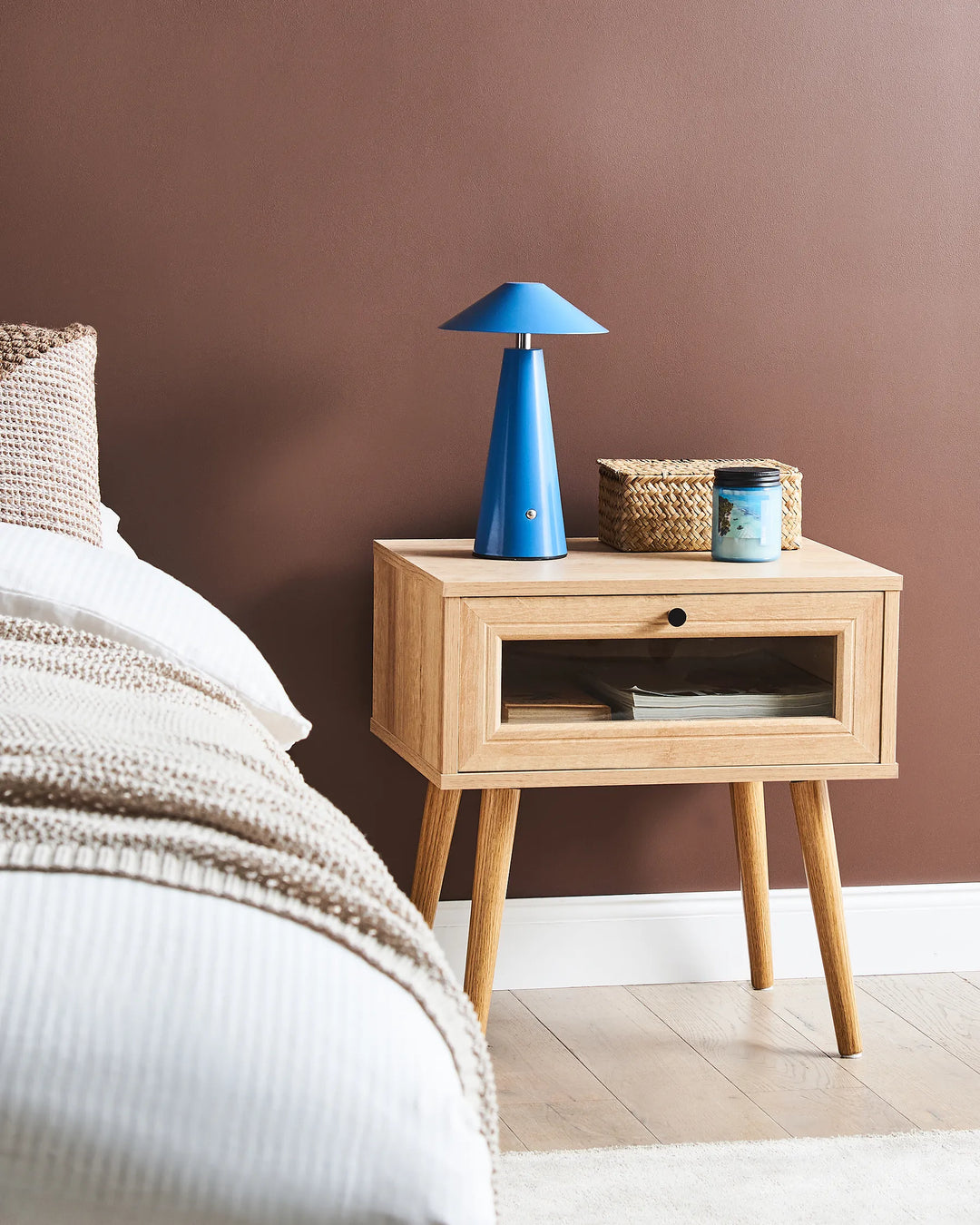 Wooden nightstand with a blue lamp and wicker basket against a brown wall.