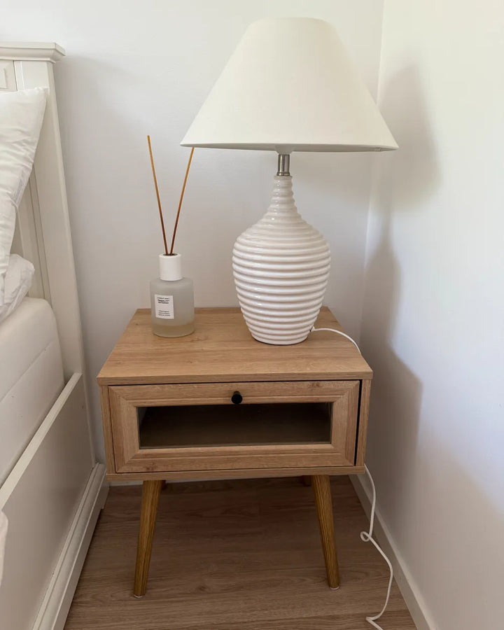 A mid-century modern bedside table made of light oak wood with tapered legs, featuring a single drawer with a black knob and an open shelf. It is styled in a bedroom corner with a white lamp, a reed diffuser, and a white bed frame against a white wall.