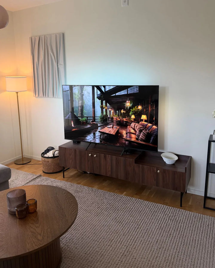 A large flat-screen television displaying a cozy living room scene, set on a dark wood media console in a modern living room with a beige rug, round coffee table, and floor lamp.
