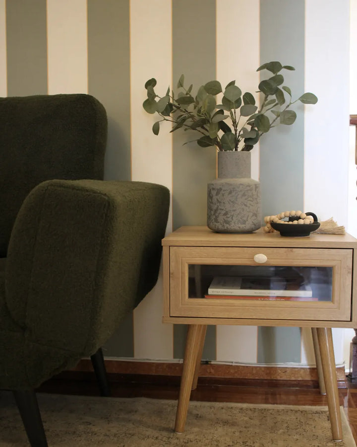 A light wood mid-century modern side table with a glass-front drawer, styled with a gray textured vase of eucalyptus and a small black bowl, next to an olive green armchair against a striped wall.