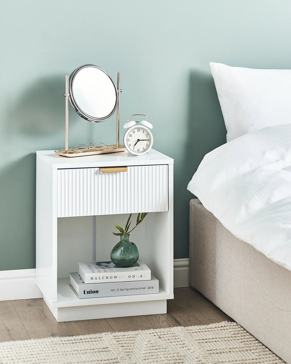 White nightstand with mirror, alarm clock, and books next to a bed with white bedding.