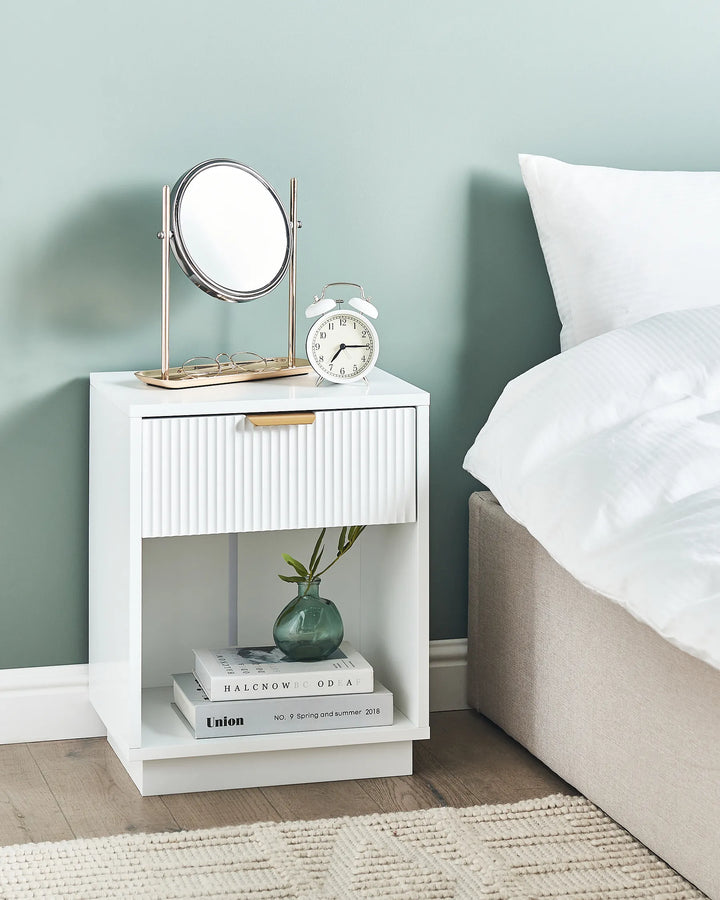 White nightstand with mirror, alarm clock, and books next to a bed with white bedding.