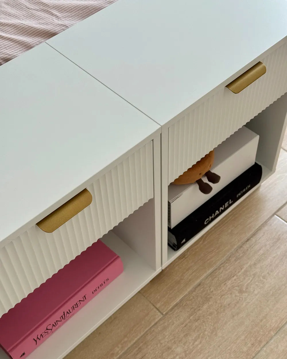 A white ribbed storage cabinet with gold handles and open cubbies, shown in a bedroom setting next to a bed with pink bedding and light wood flooring. A pink Yves Saint Laurent book and a black Chanel box are stored inside.