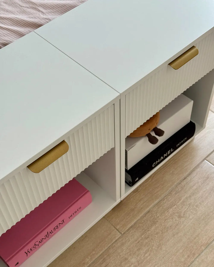 A white ribbed storage cabinet with gold handles and open cubbies, shown in a bedroom setting next to a bed with pink bedding and light wood flooring. A pink Yves Saint Laurent book and a black Chanel box are stored inside.