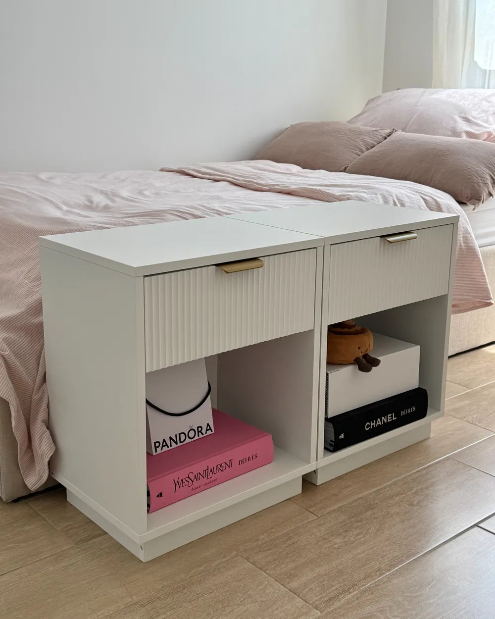 A white modern bedside table with two drawers and open shelves, styled in a bedroom next to a bed with pink bedding. The table has gold-toned metal handles and is shown holding decorative books and boxes.