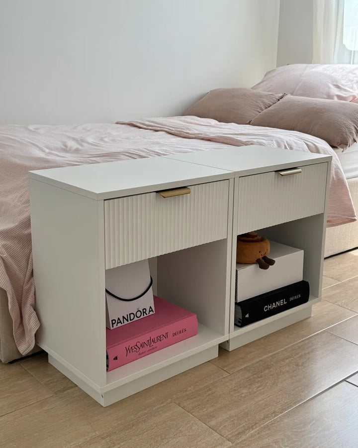 A white modern bedside table with two drawers and open shelves, styled in a bedroom next to a bed with pink bedding. The table has gold-toned metal handles and is shown holding decorative books and boxes.