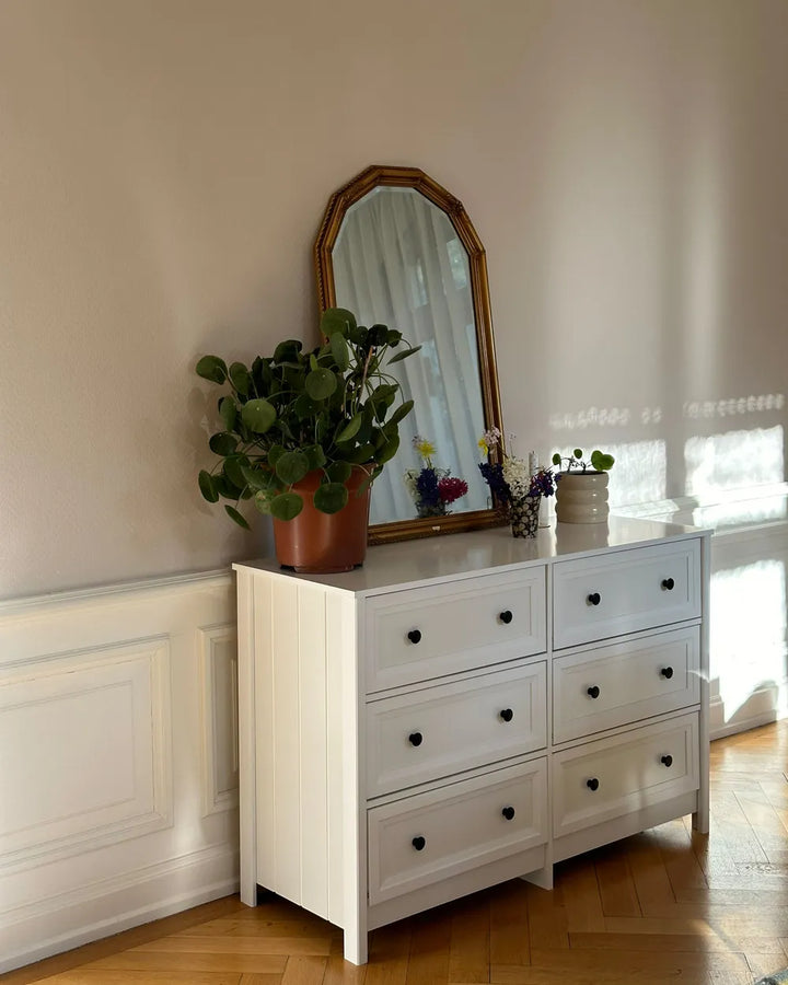 A white six-drawer dresser with black knobs in a bright room, topped with a gold-framed mirror and potted green plants.
