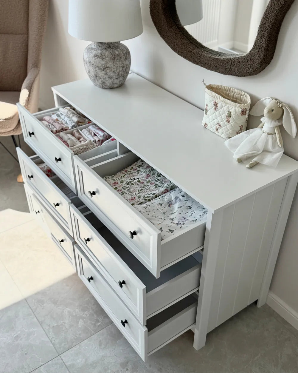 A white wooden dresser with multiple drawers, some open to show baby bedding and organizer inserts, styled in a nursery setting with a lamp, mirror, and bunny decoration.