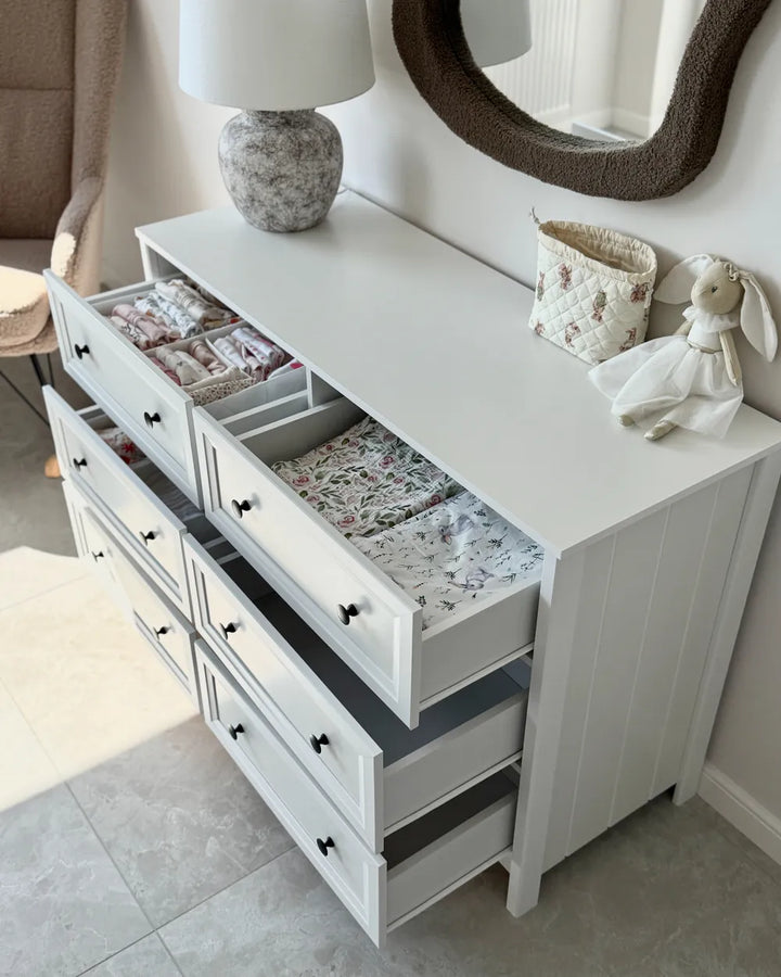 A white wooden dresser with multiple drawers, some open to show baby bedding and organizer inserts, styled in a nursery setting with a lamp, mirror, and bunny decoration.
