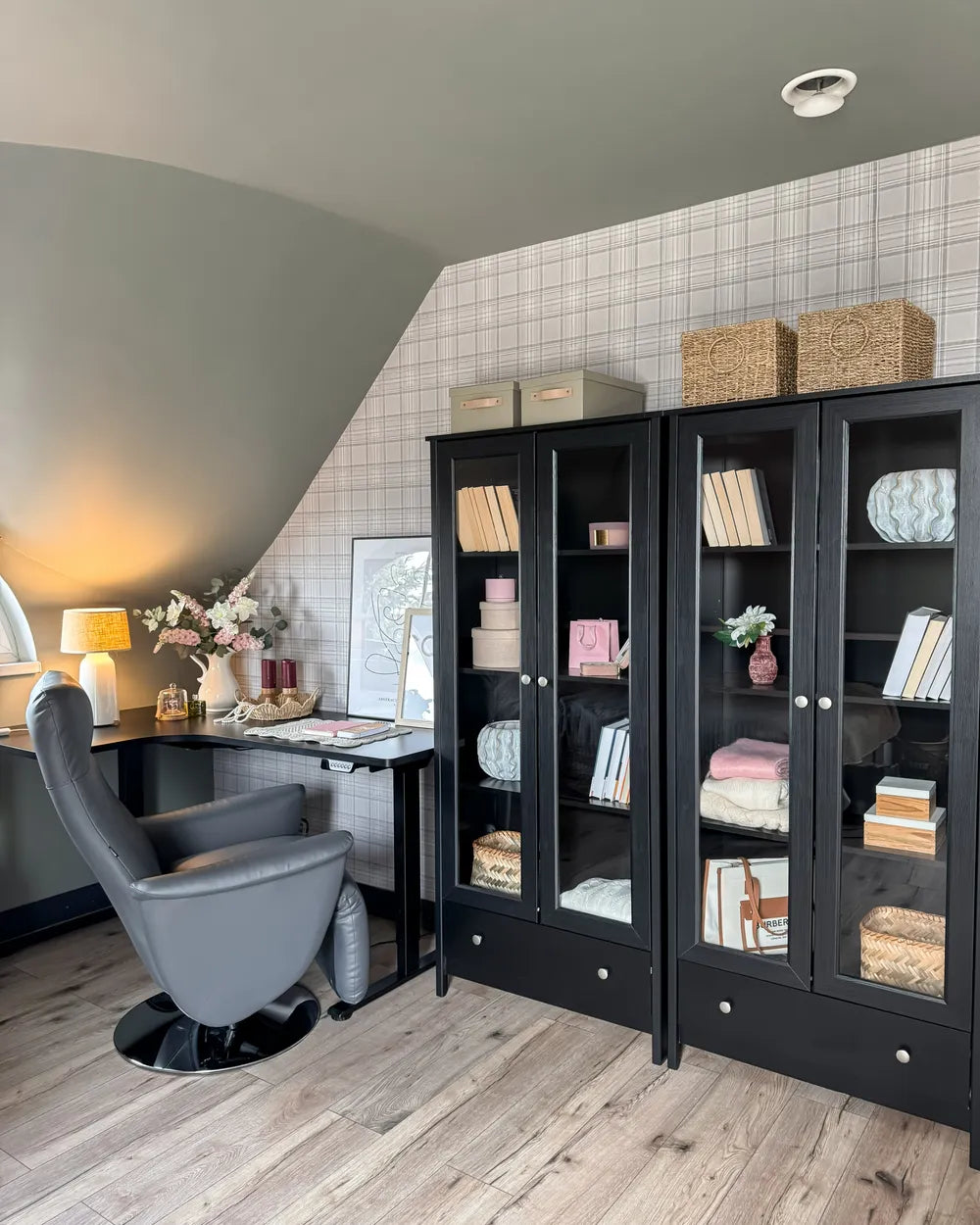 A styled home office corner featuring a large black glass-front bookcase filled with books and decor, a gray swivel chair, a desk, and a slanted ceiling with tiled wall panels.