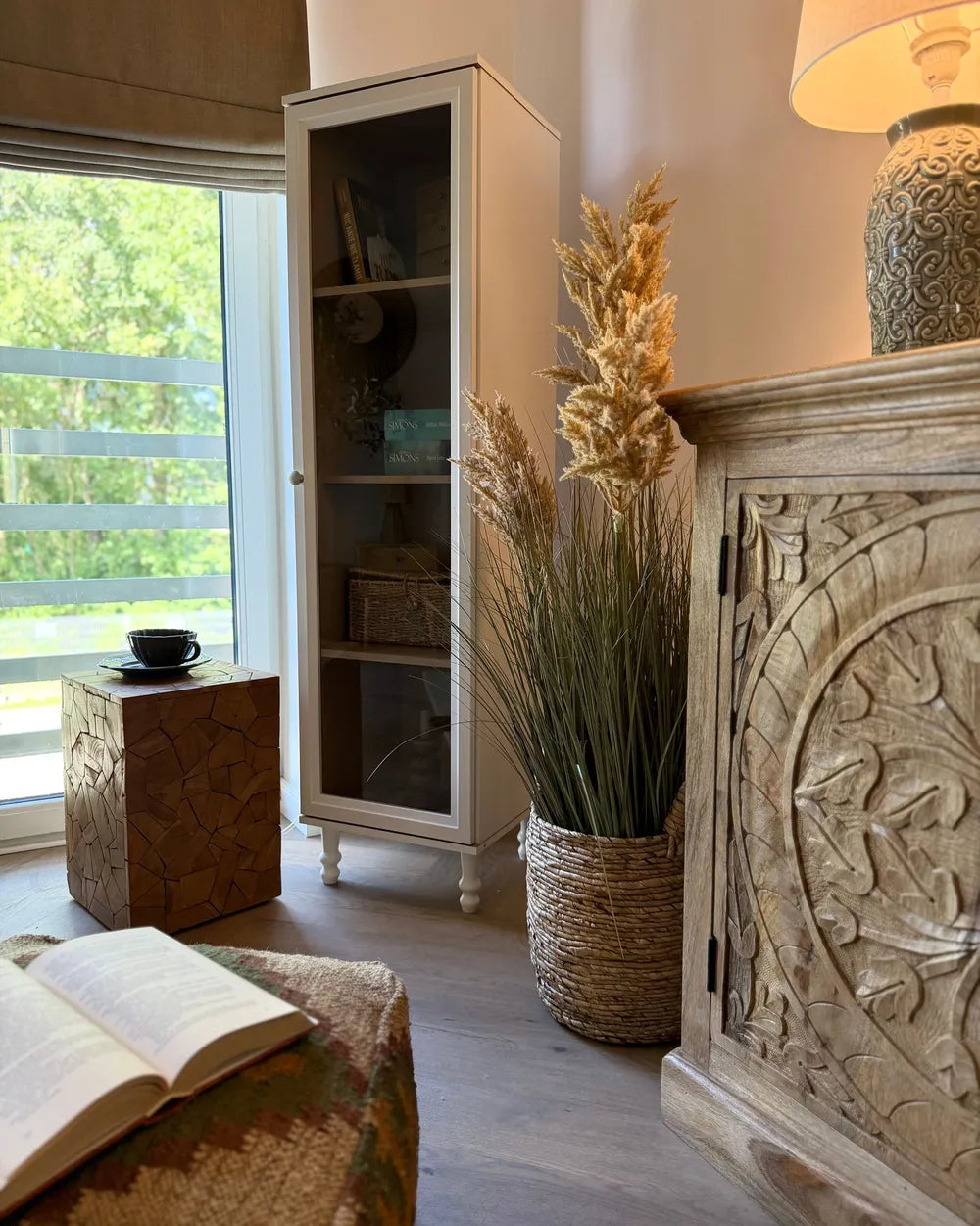 A styled interior corner featuring a tall white glass-front cabinet, a carved light wood sideboard with a lamp, a woven basket planter with dried grasses, and a window with outdoor greenery visible.