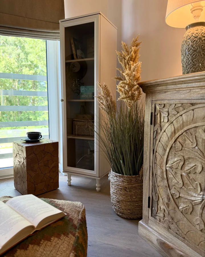 A styled interior corner featuring a tall white glass-front cabinet, a carved light wood sideboard with a lamp, a woven basket planter with dried grasses, and a window with outdoor greenery visible.
