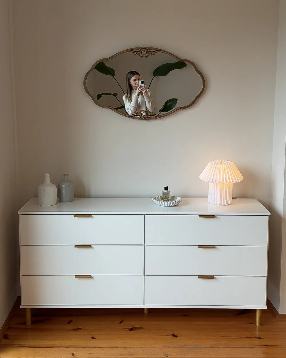 A white six-drawer dresser with gold-tone handles and legs, styled in a room with a wooden floor, a lit pleated table lamp, decorative bottles, and a decorative wall mirror.