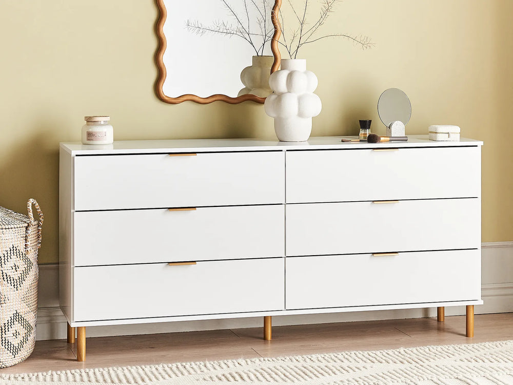 A white six-drawer dresser with slim gold-toned metal handles and tapered wooden legs, styled in a bedroom setting against a beige wall with decorative items on top.