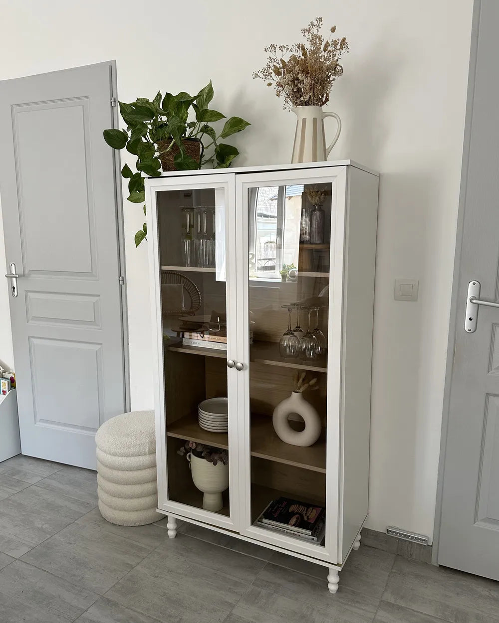 A tall white glass-front cabinet with open shelving stands in a bright room, styled with dishes, books, and dried flowers on top, next to a potted green plant and a round white pouf.