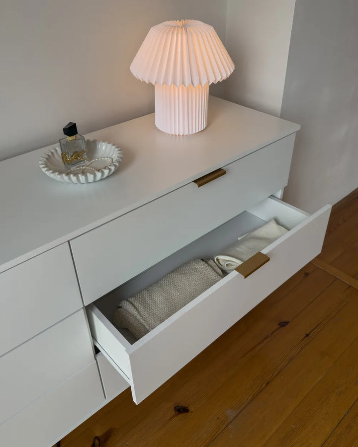 A white wooden dresser with a partially open drawer, styled with a pleated white table lamp and a small decorative tray on top, set against a neutral wall and hardwood floor.