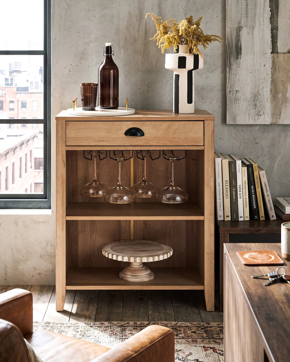 A light natural wood side cabinet with one drawer and two open shelves, styled in a loft apartment setting with wine glasses on the rack, a bottle, and a vase on top.