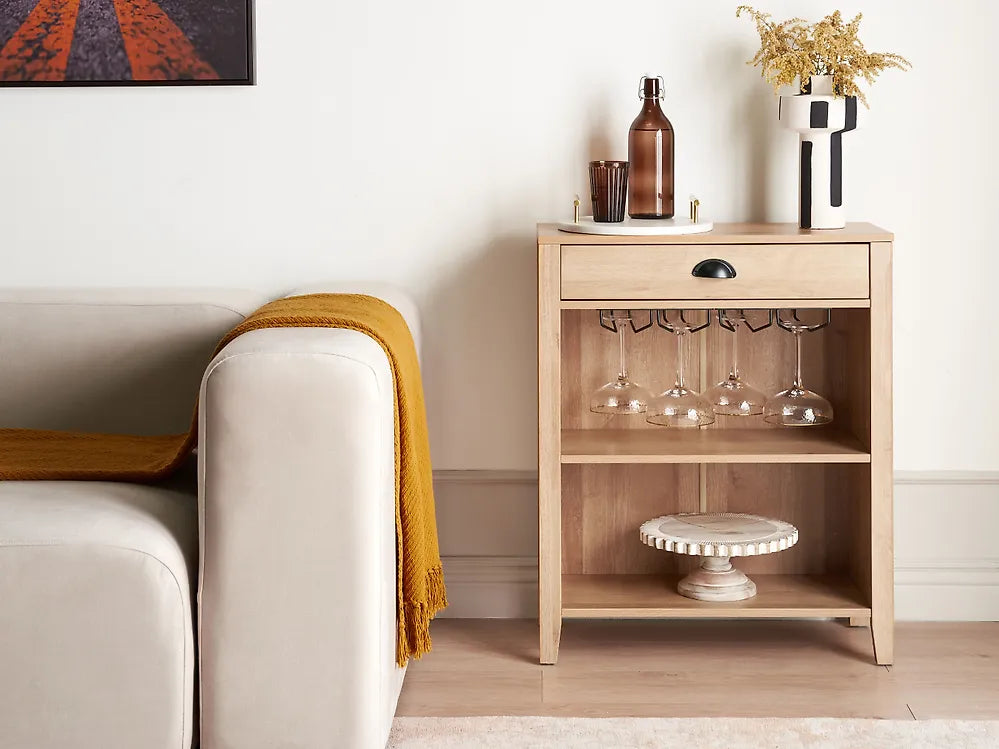 A light natural wood side table with a single drawer and two open shelves, styled in a living room next to a cream sofa with a mustard yellow throw blanket. The table is decorated with a brown glass bottle, a small candle holder, wine glasses on a rack, and a white vase with dried flowers.