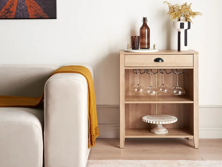 A light natural wood side table with a single drawer and two open shelves, styled in a living room next to a cream sofa with a mustard yellow throw blanket. The table is decorated with a brown glass bottle, a small candle holder, wine glasses on a rack, and a white vase with dried flowers.