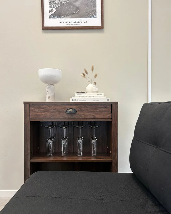 A dark brown wooden wine cabinet with a single drawer and open lower shelf holding four wine glasses, styled in a living room setting with a dark gray sofa in the foreground and a framed print on the beige wall behind.