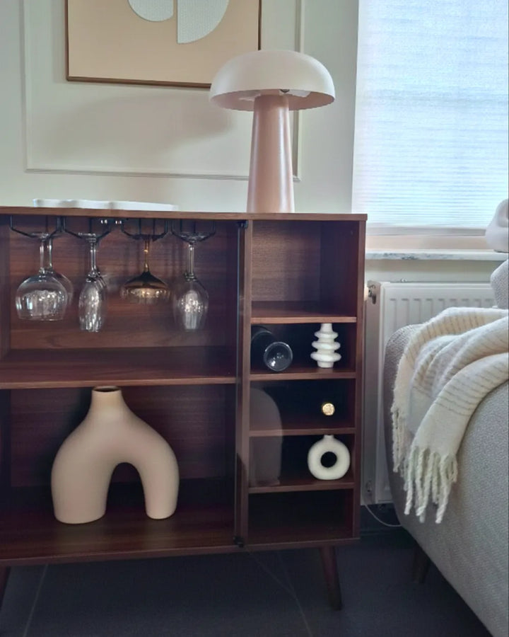 A mid-century modern wooden sideboard cabinet with open shelves, glassware hooks, and a matching mushroom-shaped table lamp on top, shown in a styled living room setting next to a sofa.