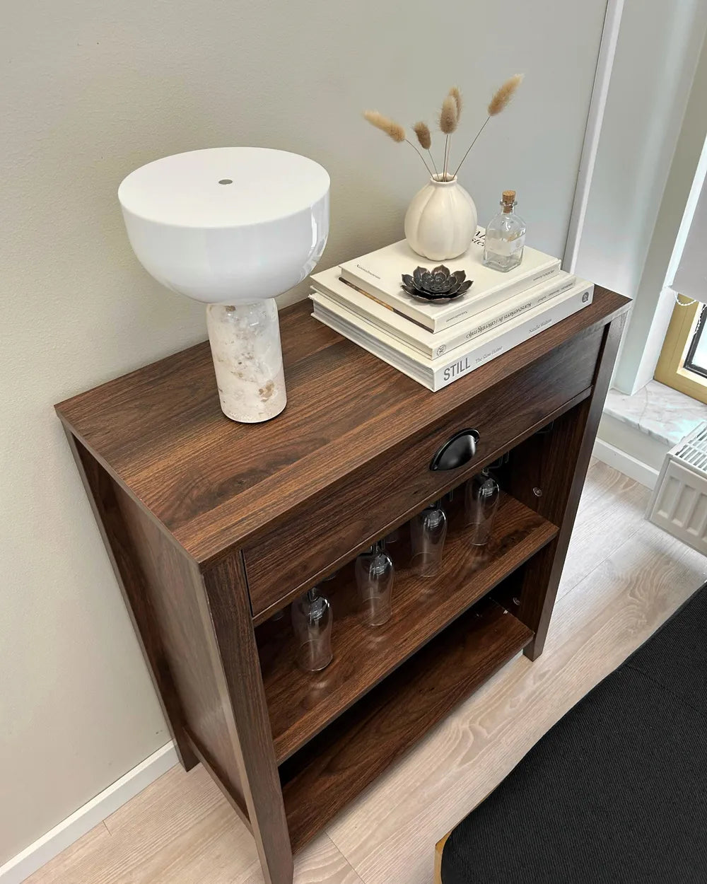 A dark brown wooden sideboard cabinet with a single drawer and two open shelves, styled with a white ceramic lamp, books, a small vase, and clear drinking glasses on the shelves, set in a home interior.