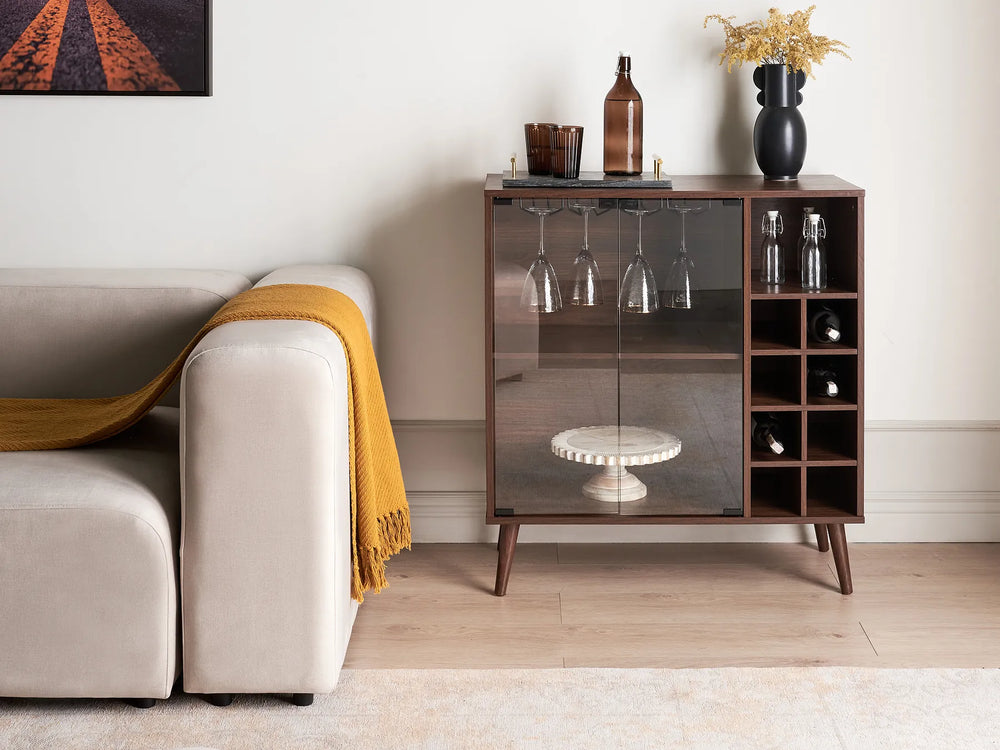 A mid-century modern wooden bar cart with glass doors and wine bottle storage, styled in a living room next to a beige sofa with a yellow throw blanket.