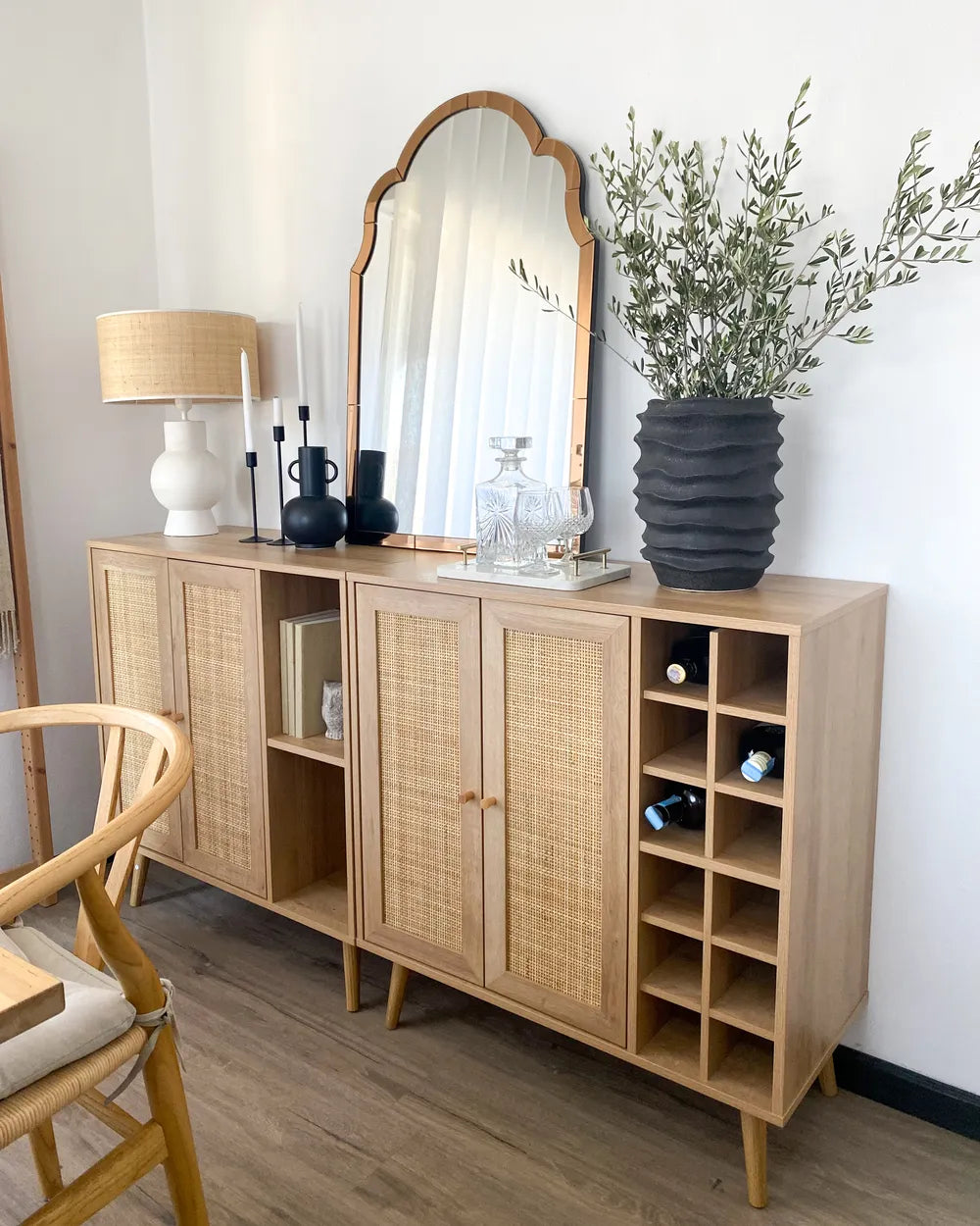 A light natural wood sideboard cabinet with woven cane doors, styled in a bright room with a large arched mirror, a table lamp, candles, and a black ribbed vase holding green branches.
