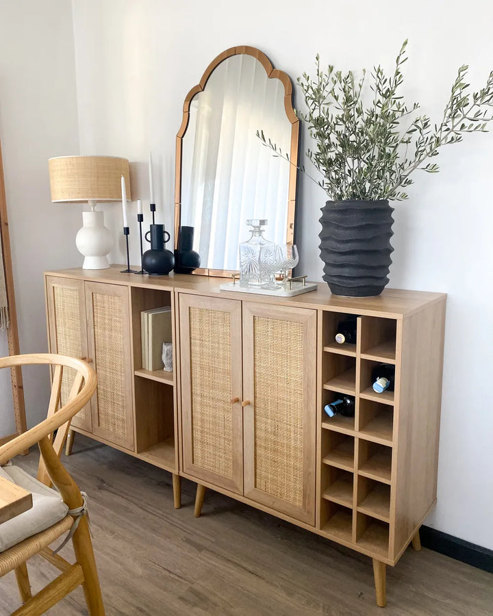 A light natural wood sideboard cabinet with woven cane doors, styled in a bright room with a large arched mirror, a table lamp, candles, and a black ribbed vase holding green branches.