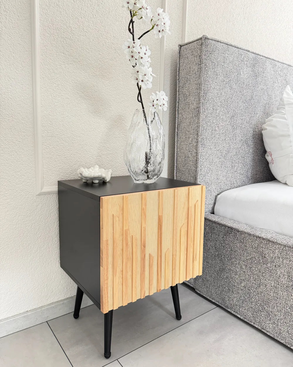 A modern bedside table with a matte black side panel and a slatted light wood front, styled next to a bed with a gray upholstered headboard in a bedroom setting.