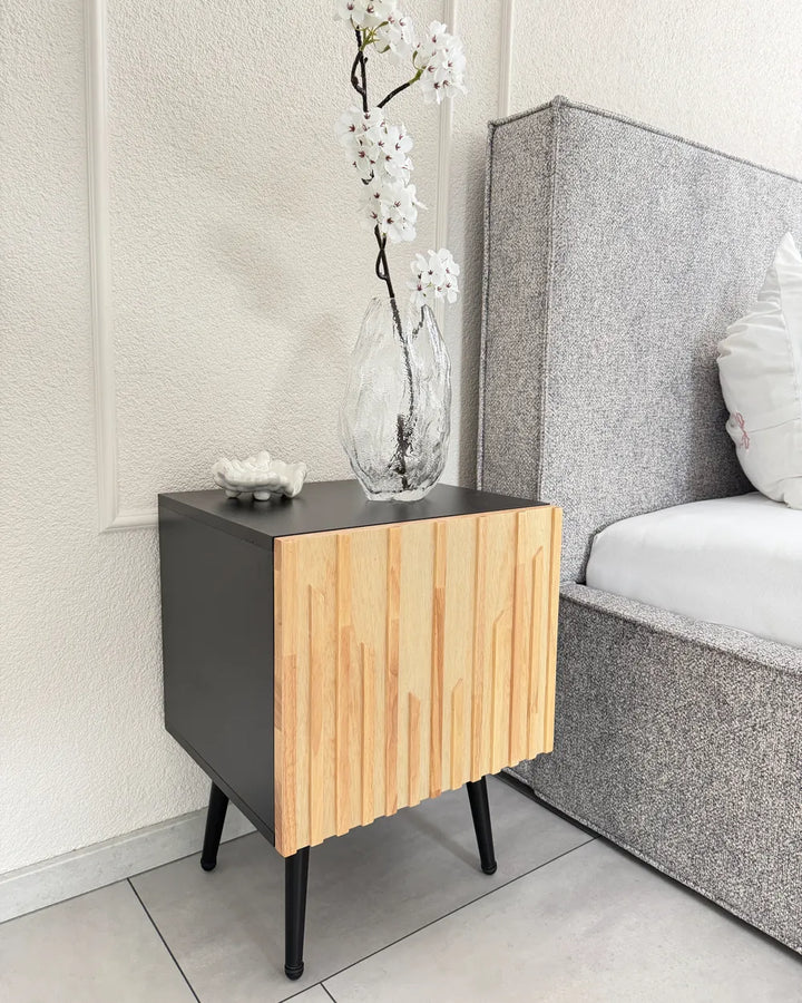 A modern bedside table with a matte black side panel and a slatted light wood front, styled next to a bed with a gray upholstered headboard in a bedroom setting.