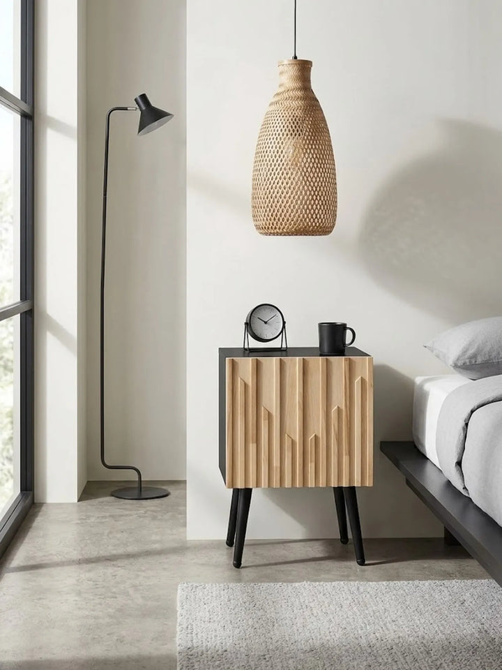 A modern bedroom scene featuring a light wood nightstand with black legs and a black top, styled with a small clock and mug. A black floor lamp stands to the left next to a window, and a woven pendant light hangs above.