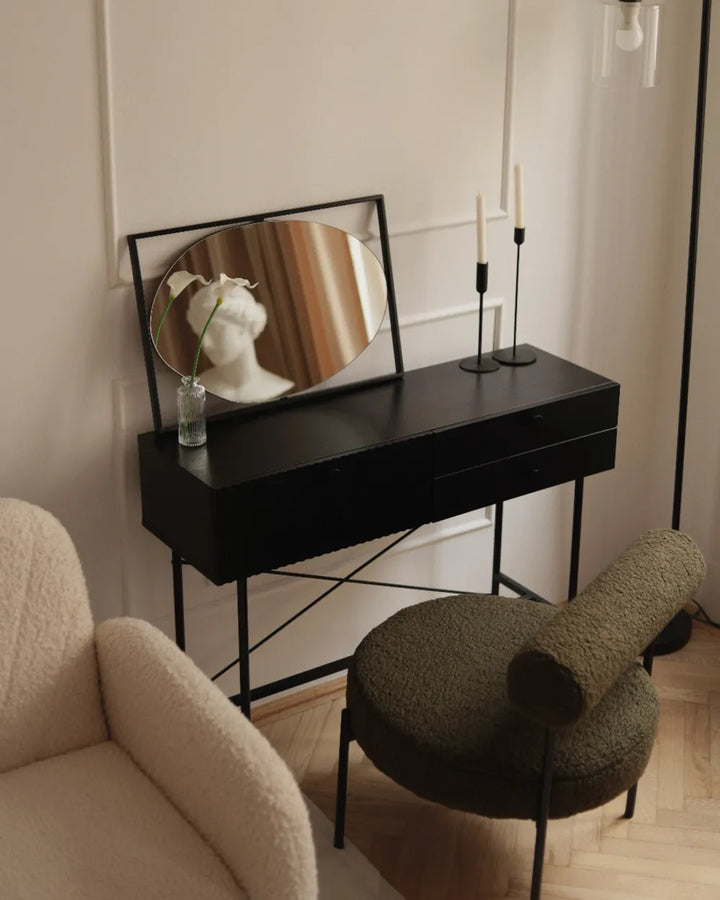 A black console table with thin metal legs in a styled room, featuring a black-framed oval mirror, a small glass vase with a white flower, and two black candlesticks. A textured olive green boucle stool is in front, and part of a cream boucle armchair is visible on the left.