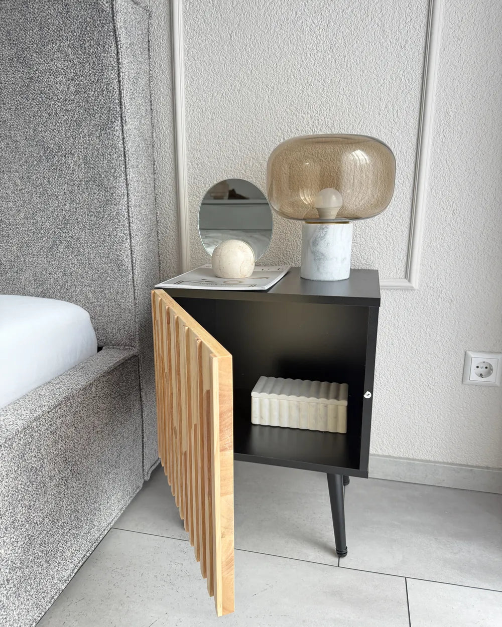 A modern black bedside cabinet with a light wood slatted door panel, styled in a bedroom next to a bed with a gray upholstered headboard. The cabinet is open, showing an interior shelf with a white ribbed box, and is topped with a round mirror, a white sphere, and a translucent amber glass table lamp.