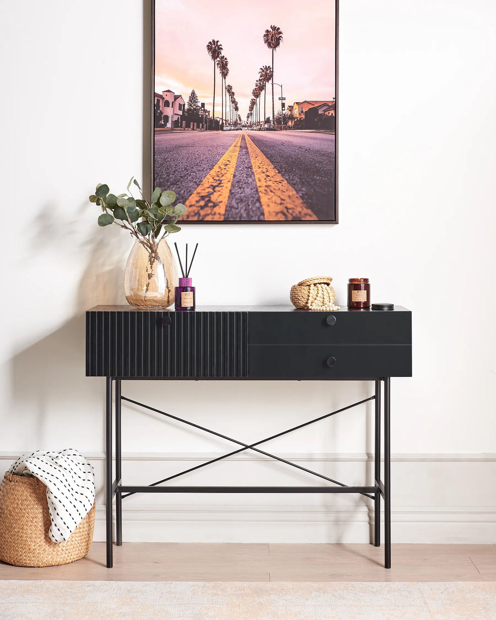 A black console table with a ribbed front and open crisscross metal legs, styled in a bright room with a framed palm tree print on the wall, a glass vase with greenery, and small decor items on top.