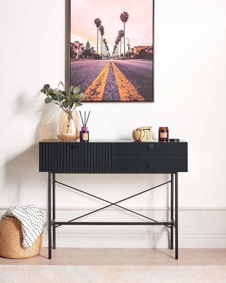 A black console table with a ribbed front and open crisscross metal legs, styled in a bright room with a framed palm tree print on the wall, a glass vase with greenery, and small decor items on top.