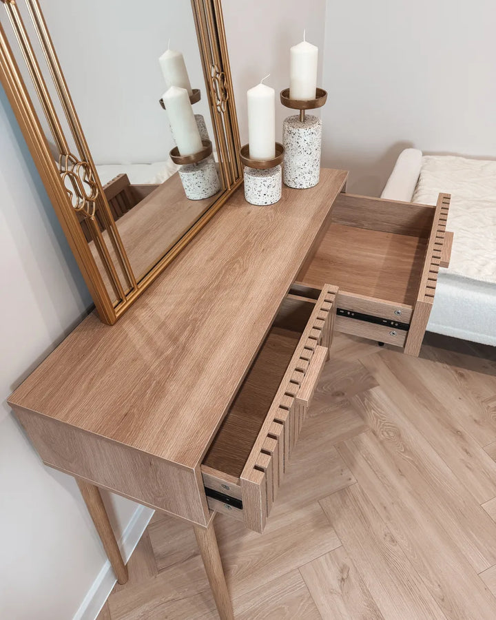 A light oak wood vanity table with two open drawers and a large gold-framed mirror on top, styled with white candles in a bedroom setting.