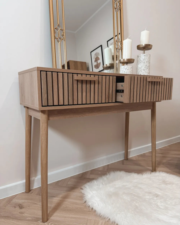 A mid-century modern wooden console table with a slatted front and tapered legs, styled in a room setting with a gold-framed mirror, candles, and a white faux fur rug on a light wood floor.